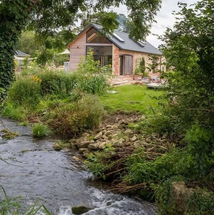 A photograph of a building with a steep pitched roof, skylights, and large glass doors, featuring both timber cladding and brickwork. It is situated directly behind a lush, overgrown green riverbank with a shallow rocky stream flowing in the foreground.