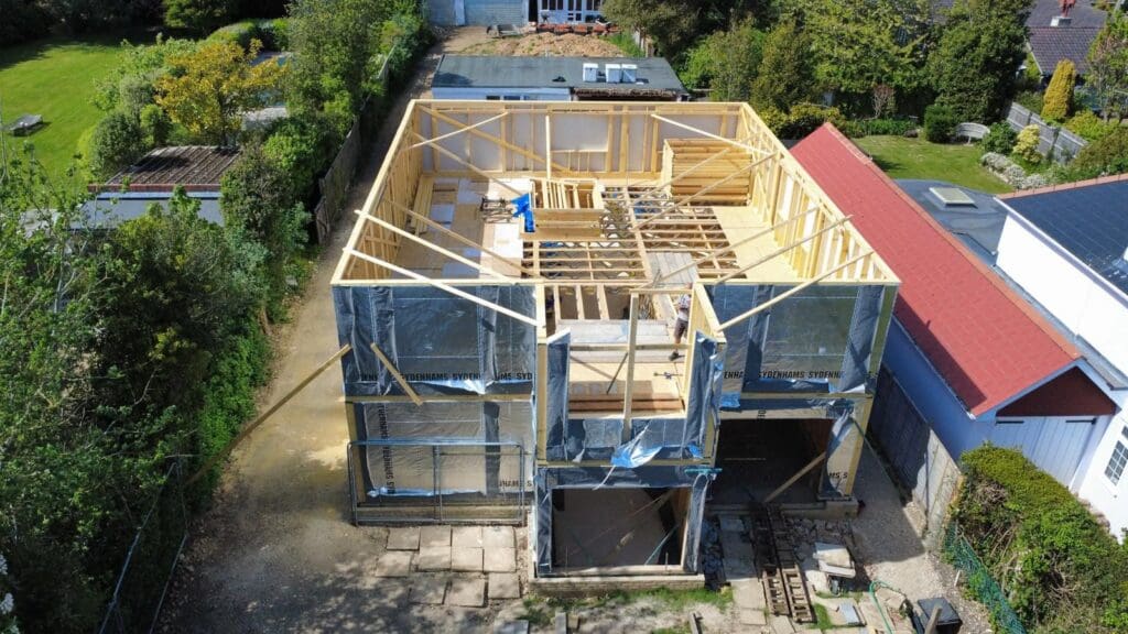 An aerial view of a two-story house under construction. The exposed timber framing of the upper walls and floor joists is visible, while the lower exterior walls are partially wrapped in a silver building membrane printed with the word "SYDENHAMS". Neighboring residential houses and green gardens surround the construction site.